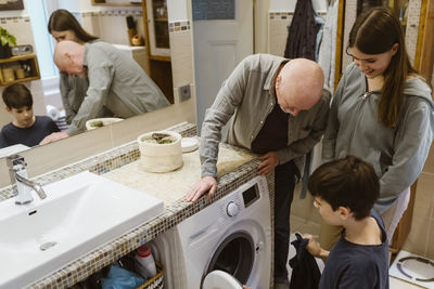 Senior man teaching grandchildren to use washing machine in bathroom at home