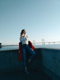 Full length of young woman sitting on retaining wall against clear sky