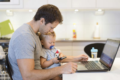 Father with laptop looking at baby boy using mobile phone at table