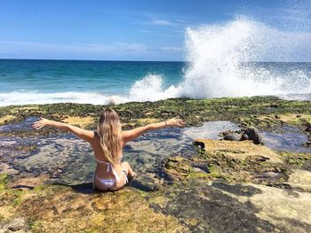 Woman with arms outstretched sitting in front of waves splashing on rock formation