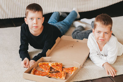 Two boys lie on the floor eating pepperoni pizza from a box. vertical frame. 