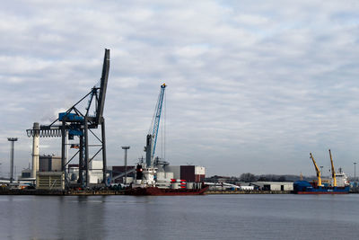 Cranes at commercial dock by river against cloudy sky
