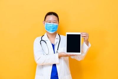 Portrait of smiling doctor showing digital tablet standing against yellow background