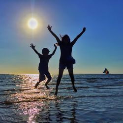 Full length of silhouette siblings jumping with arms raised against sea