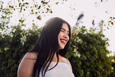 From below happy ethnic female with long black hair and red lips smiling with closed eyes while standing on blurred background of lush bush on sunny day in park