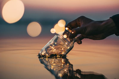 Cropped image of hand putting glass jar in sea during sunset