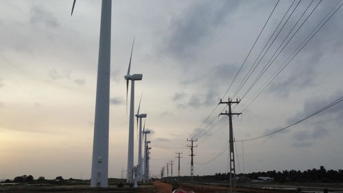 Low angle view of electricity pylons and wind turbines against cloudy sky