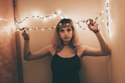 Portrait of young woman with illuminated string lights standing against wall