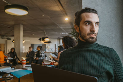 Thoughtful young bearded man sitting with laptop in university cafeteria