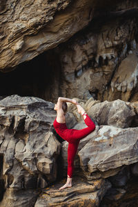 Side view of young woman in red sportswear doing yoga on lord of the dance pose standing on one leg while outstretching different leg on rough rock among mountains