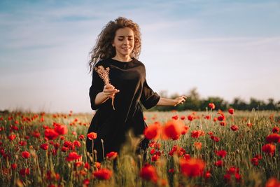 Young woman standing by poppy flowers on field against sky