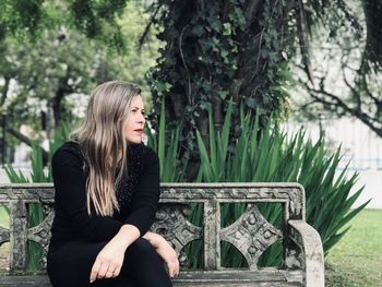 Young woman looking away while sitting on seat against trees