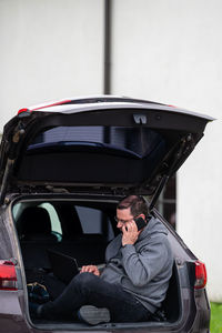 A man sitting in the open trunk of a car and working on a laptop, technology to work remotely