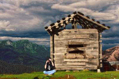 Woman sitting by log cabin on field against cloudy sky