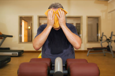 Man wiping face with towel while sitting in gym