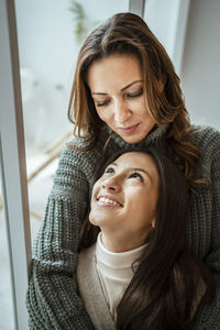 Beautiful mother and daughter looking at each other while sitting by window at home