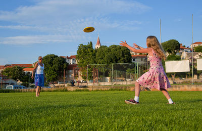 Woman and girl playing with plastic disc on grass against sky