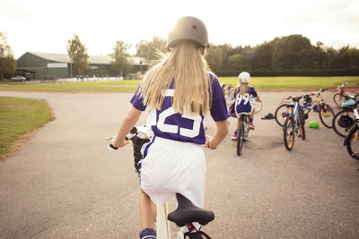 Rear view of girls cycling on footpath by soccer field against sky