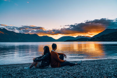 Woman sitting by lake against sky during sunset