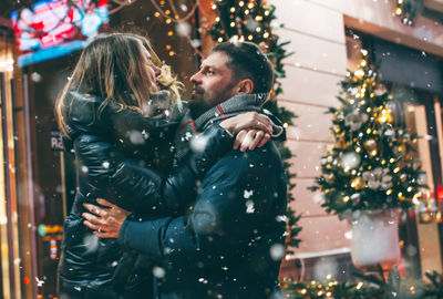 Couple romancing while standing against buildings at night during winter in city