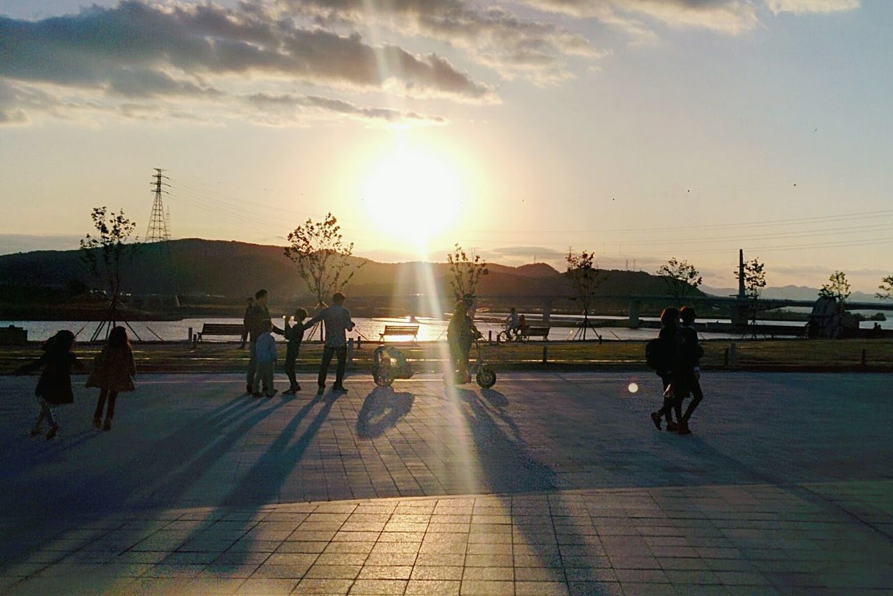 sky, sunset, lifestyles, men, water, sunlight, leisure activity, person, sun, walking, sea, cloud - sky, silhouette, cloud, full length, built structure, horizon over water, medium group of people, street light