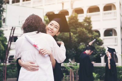 Cheerful woman in graduation gown embracing mother while standing outdoors