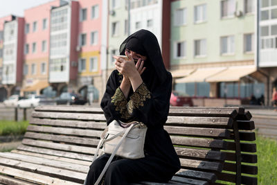 Woman paints her eyes while sitting on a bench on the street.