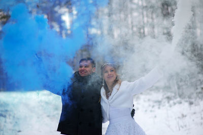 Smiling wedding couple holding distress flares while standing at forest during winter