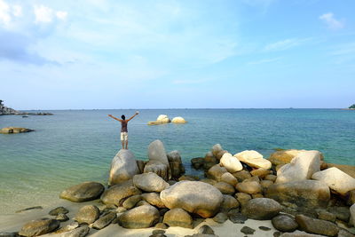 Rear view of man with arms outstretched standing on rock by sea against sky