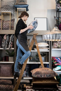 Full length of young female entrepreneur standing on ladder while arranging cushion in rack at store