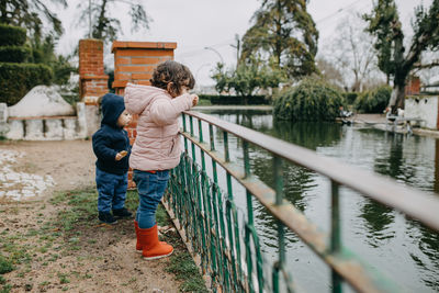 Side view of siblings standing by railing over lake against trees