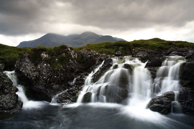 Scenic view of waterfall from mountains against cloudy sky