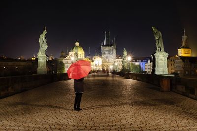 Rear view of woman with umbrella standing against illuminated buildings in city at night