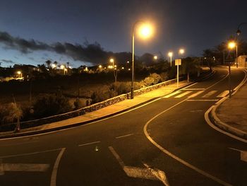 Empty road by illuminated city against sky at night