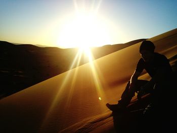 Silhouette man sitting on sand dune against sky at sahara desert during sunny day