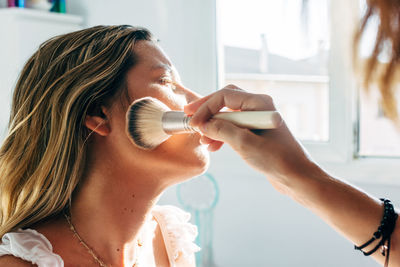 Cropped unrecognizable female makeup artist with powder and brush doing makeup to a friend at home