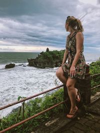 Side view of woman looking at sea while sitting on railing against cloudy sky