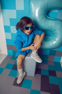 Boy child in blue clothes and a cap is sitting on a gray square celebrating his birthday. 