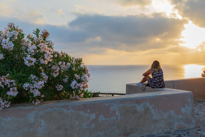 Woman sitting by sea against sky during sunset