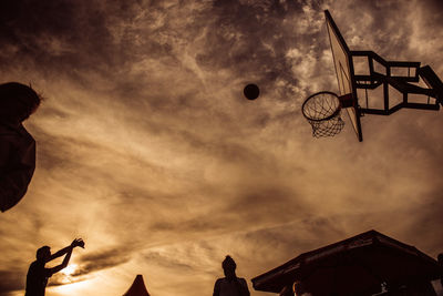 Low angle view of people playing basketball against cloudy sky during sunset