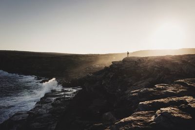 Distant view of person standing on cliff by sea against clear sky during sunset