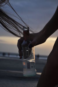 Close-up of hand holding glass bottle against sky during sunset