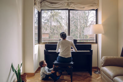 Rear view of mother and son playing piano at window