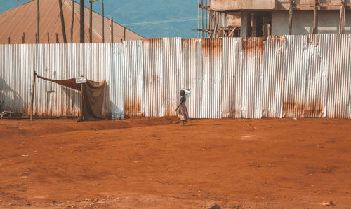 Full length of a child carrying a box on her head against a corrugated iron and brown sand