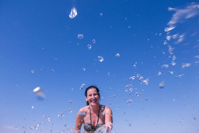 Smiling woman splashing water against clear blue sky during sunny day
