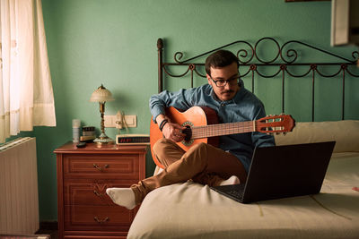 Young man practices with the guitar by looking at his laptop at home