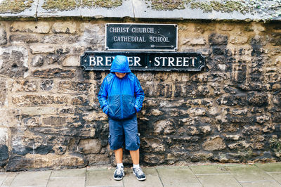 Full length of boy in hooded jacket standing against information sign on stone wall