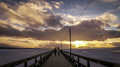 People on footbridge over snow covered field during sunset
