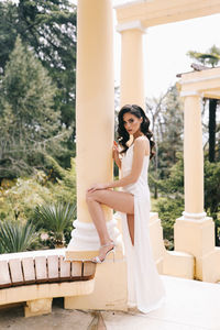 A beautiful brunette lady in an elegant wedding dress poses among the columns in the old city park