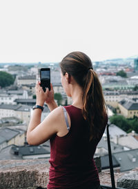 Rear view of woman photographing on mobile phone while standing in balcony against sky